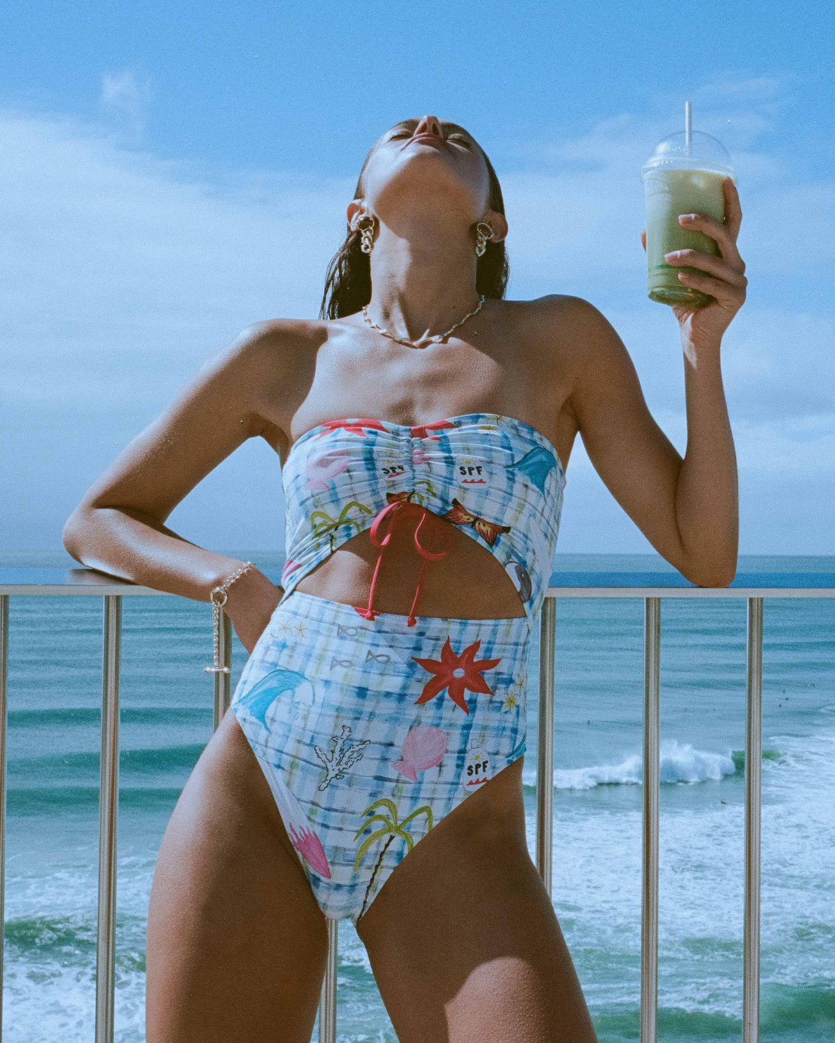 White bandeau one-piece swimsuit with blue gingham pattern and multicolored beach-themed graphics including palm trees and st