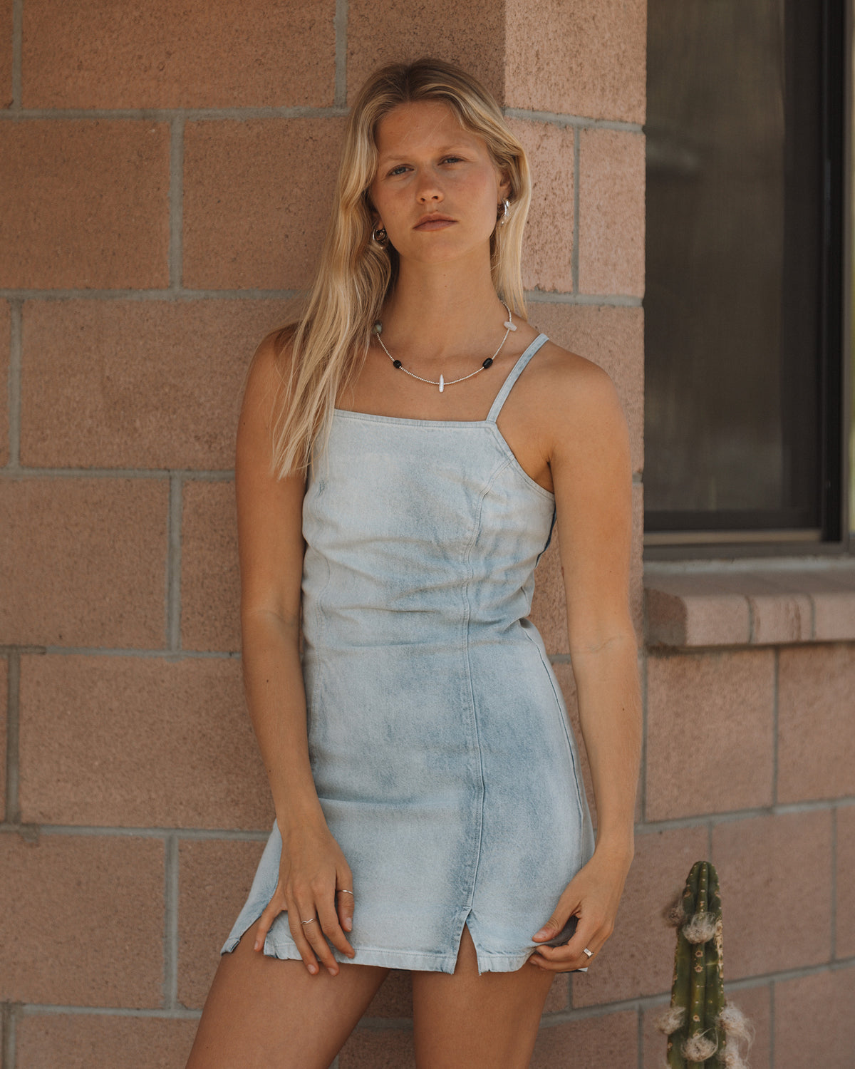 Light blue denim dress with thin straps and front slit detail on model against brick wall.