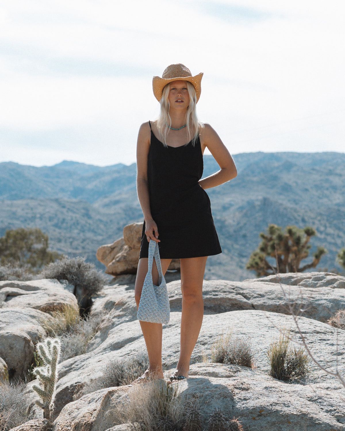 Black sleeveless slip dress with thin straps worn with a straw cowboy hat and blue shoulder bag.