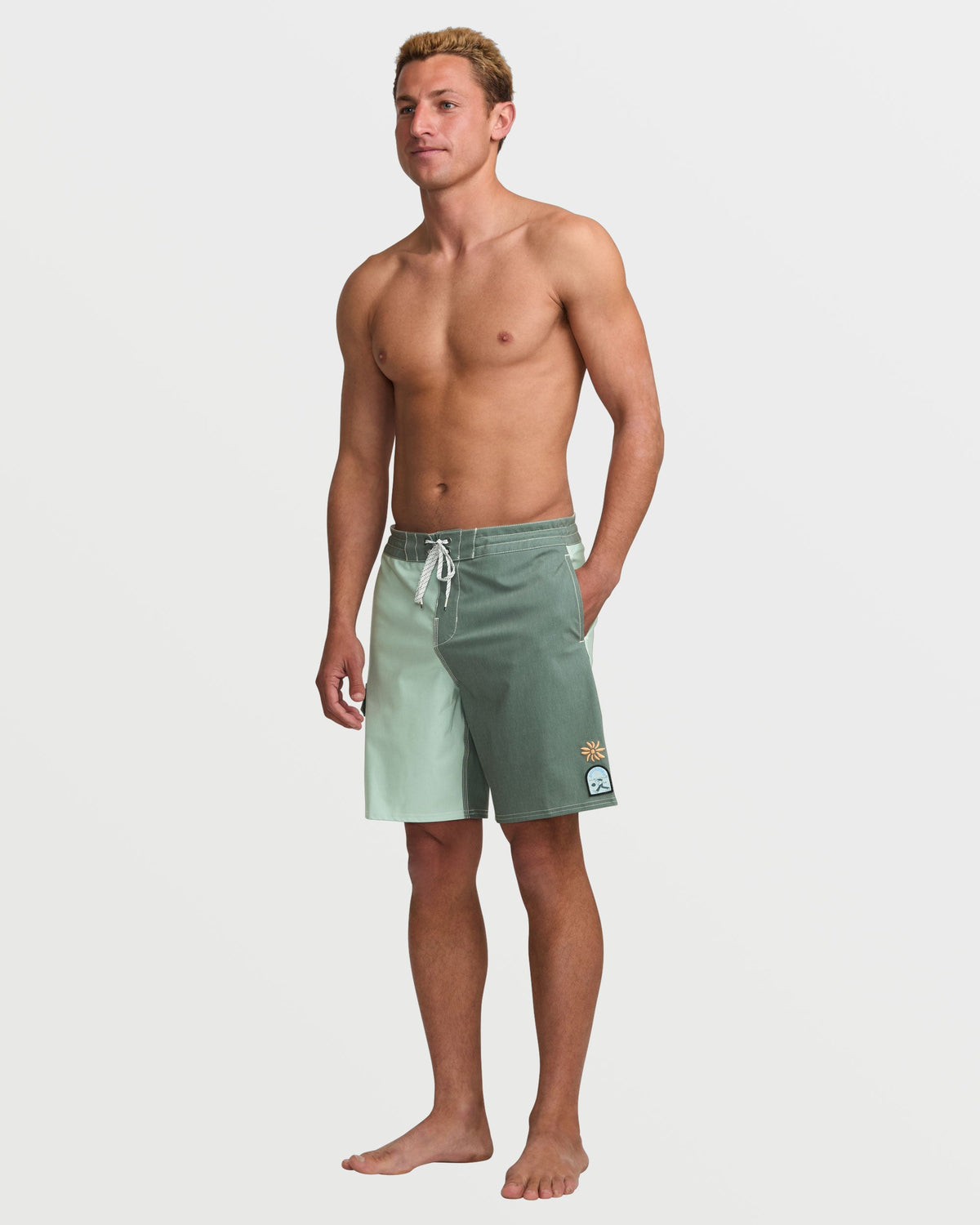 Man wearing green tea-colored boardshorts with drawstring waist and logo patch on leg.
