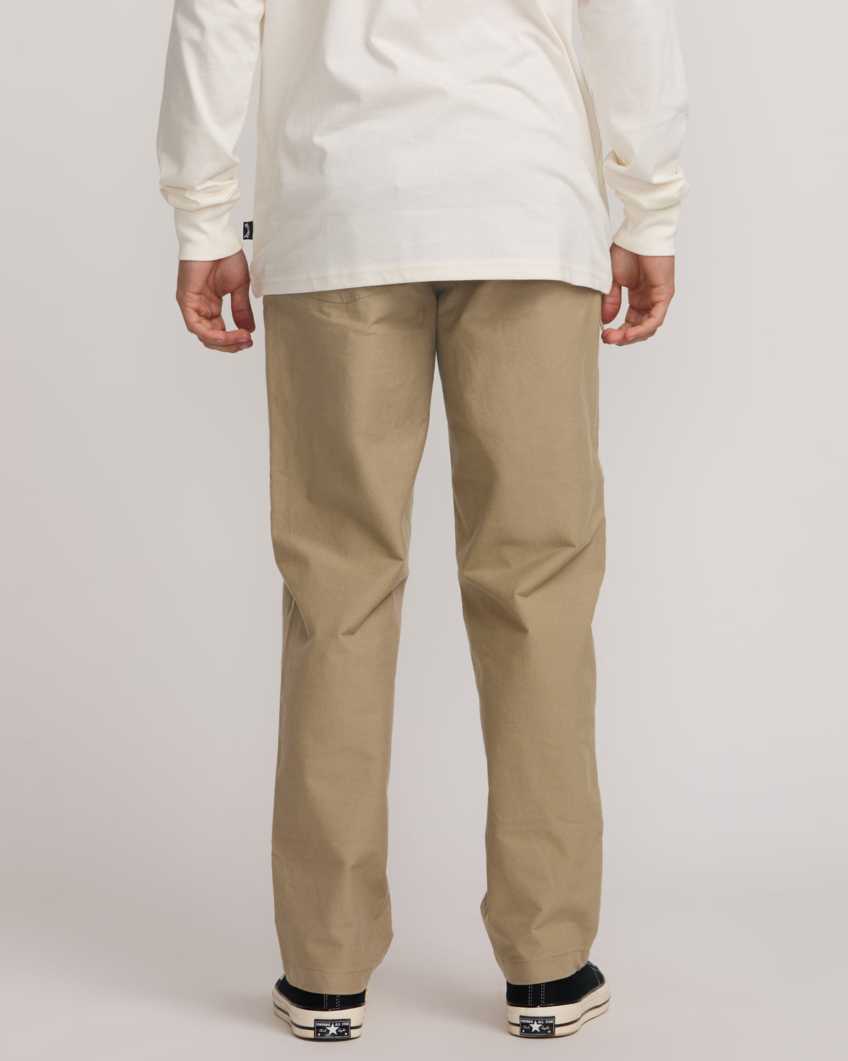 Beige flat-front pants with straight legs, shown worn with white long-sleeve shirt and black sneakers.