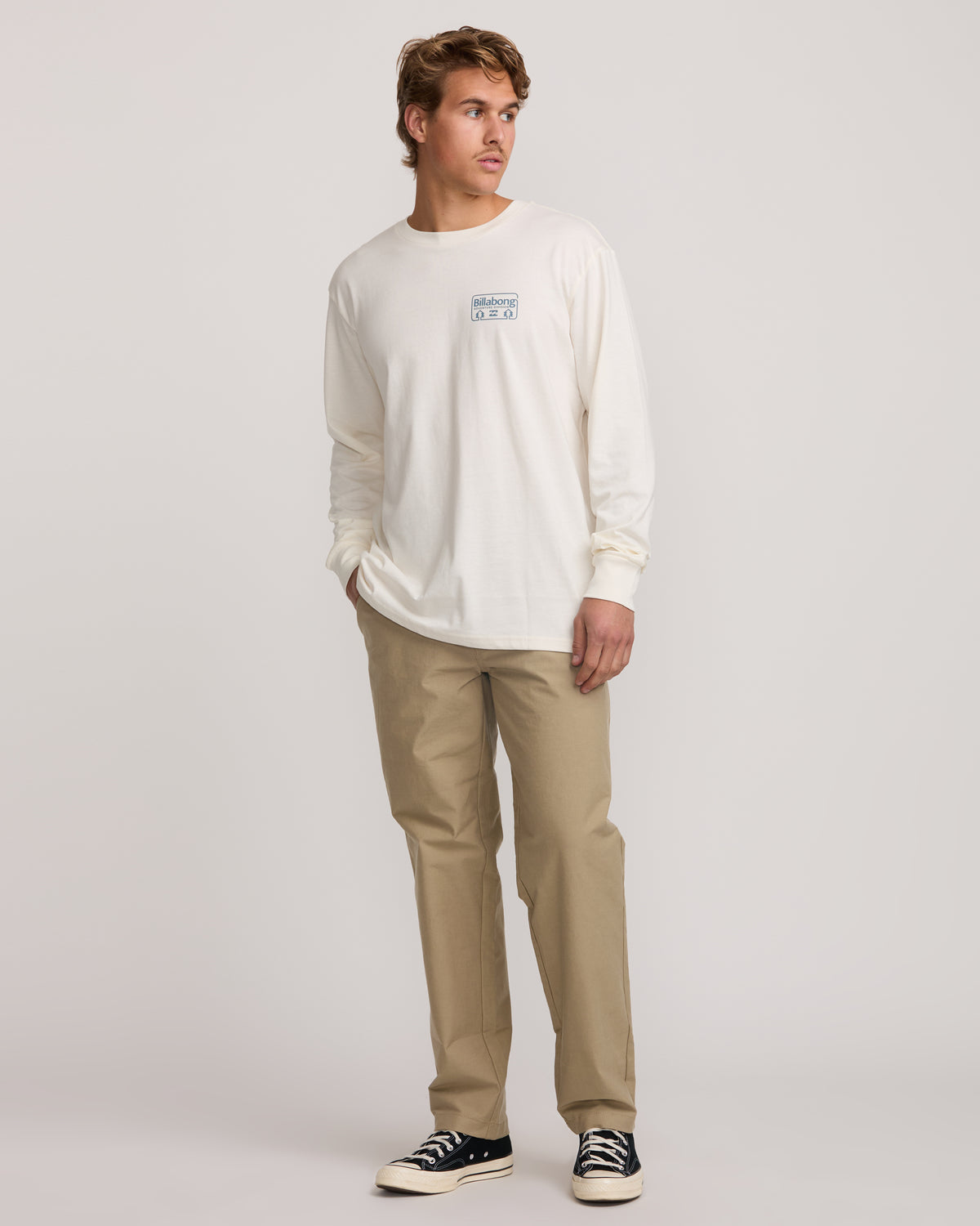 Man wearing cream long-sleeve shirt with small chest logo and gravel-colored straight-leg pants.
