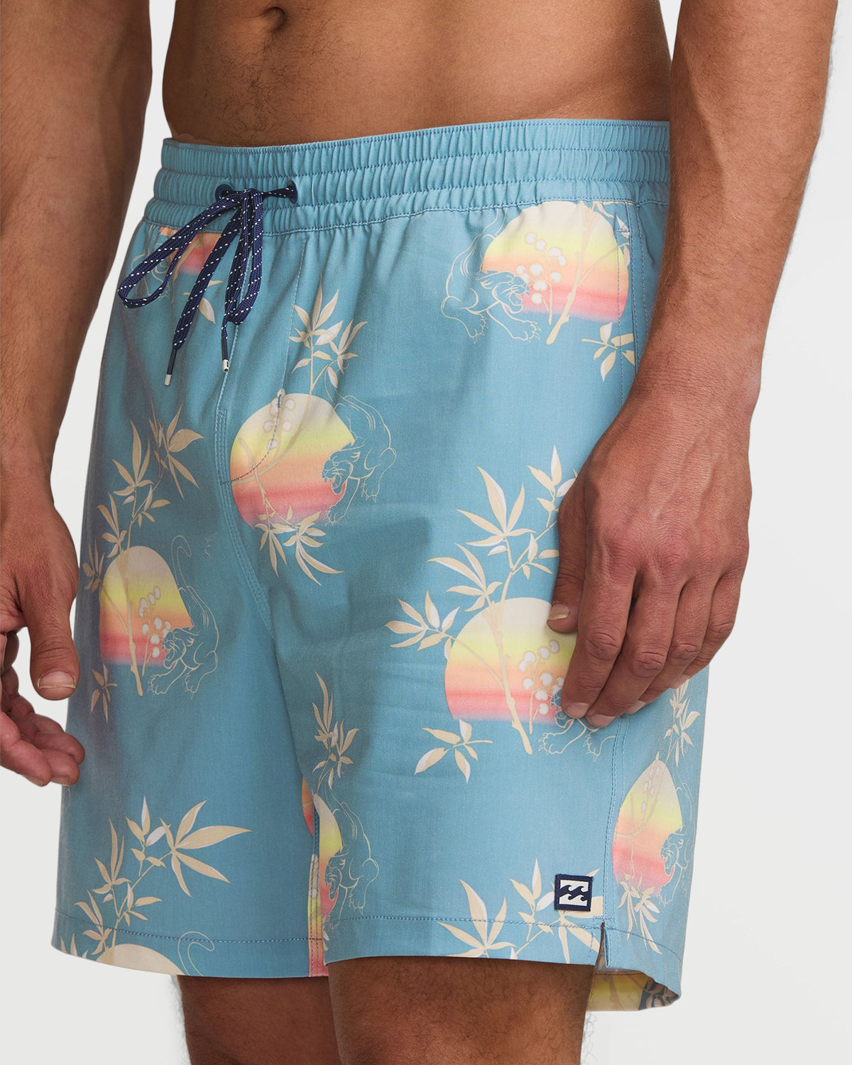 Washed blue swim trunks with sunset and palm leaf print, drawstring waist, and solid navy tie closure.