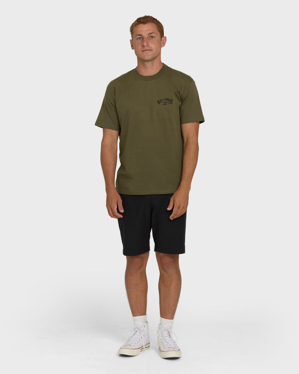 Olive green short-sleeve t-shirt with small chest logo, shown on model wearing black shorts and white sneakers.