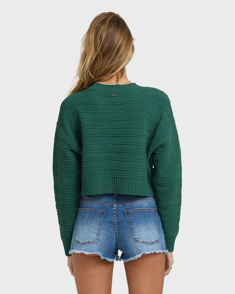 Green ribbed knit sweater with long sleeves shown from the back worn with denim shorts.