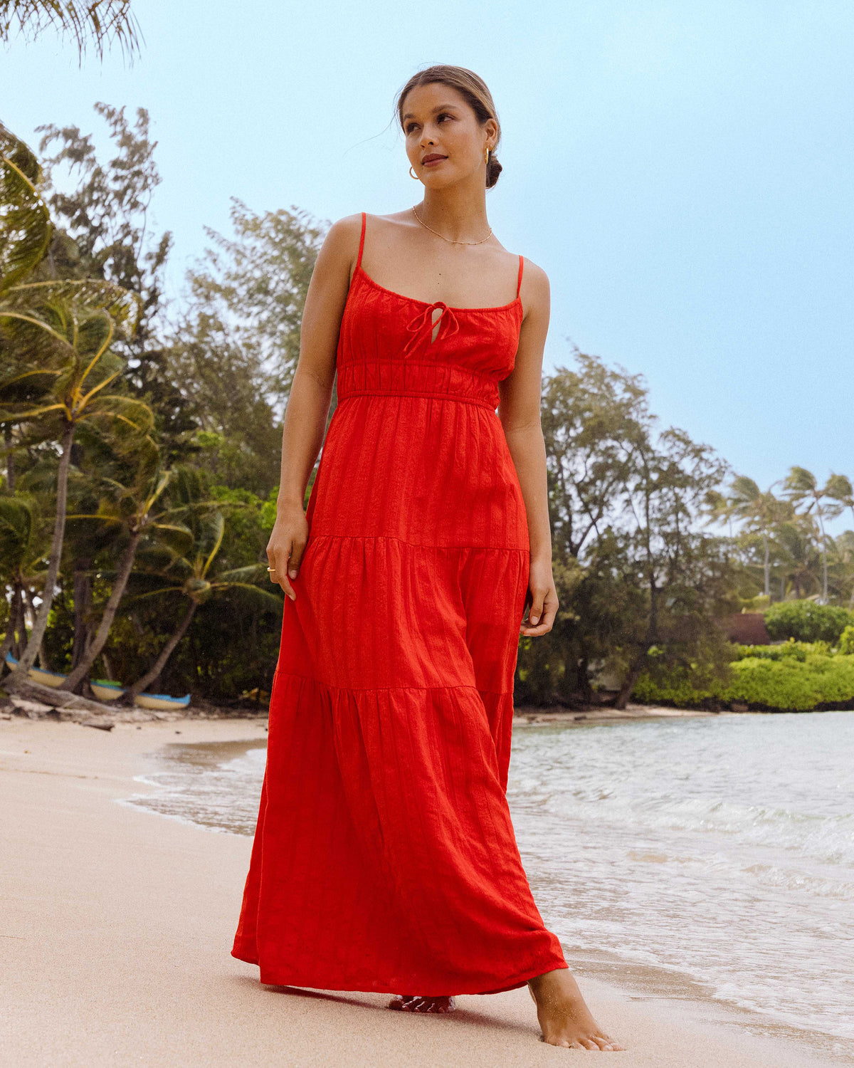 Woman wearing red sleeveless maxi dress with tiered ruching and tie-front neckline, standing on a beach.