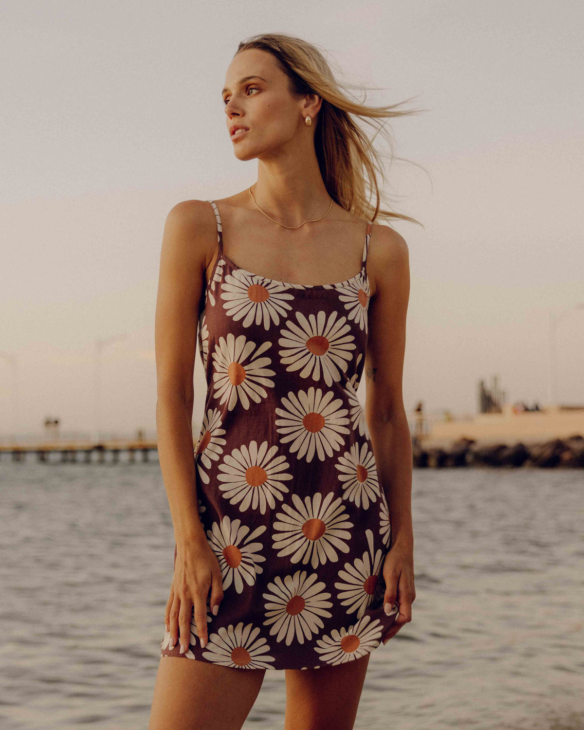Dark brown sleeveless dress with white and orange daisy floral print, thin straps, worn by woman at beach.