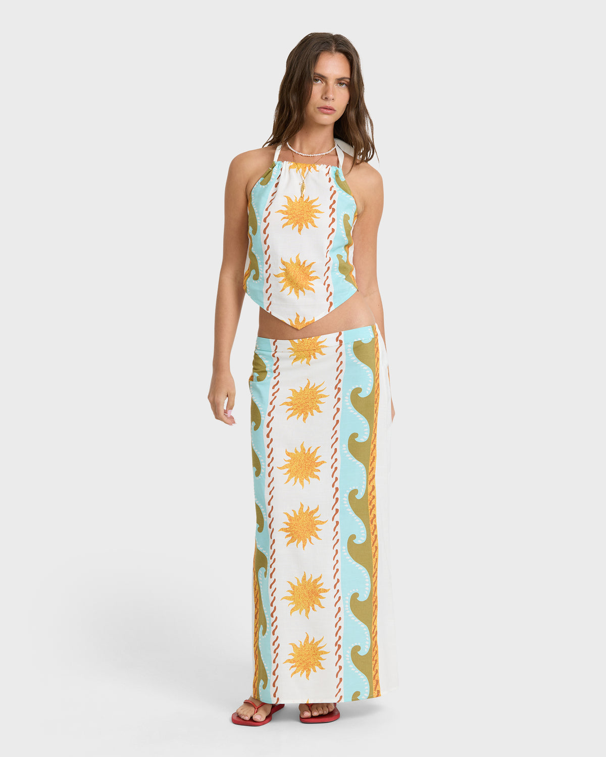 Woman wearing a two-piece outfit with a halter top and midi skirt featuring sun and wave motifs in yellow, turquoise, and whi