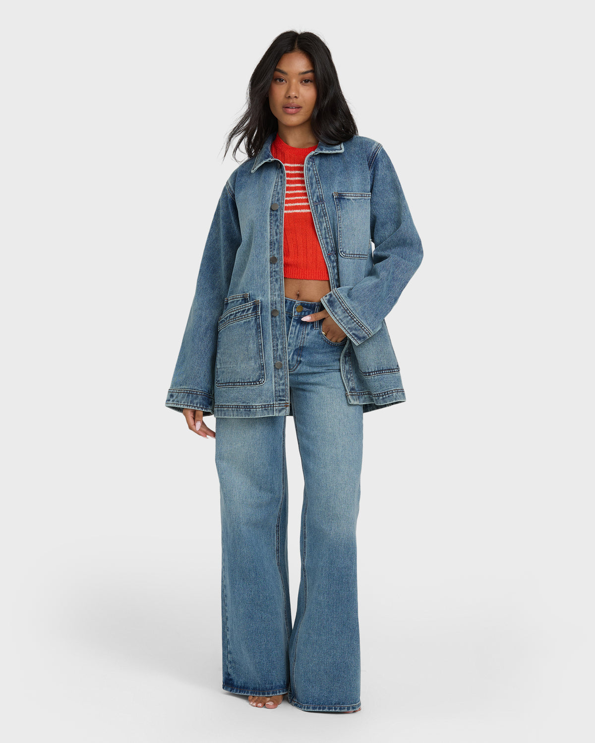 Medium-wash denim chore coat with button front, collar, and chest pocket worn over red striped top and light-wash jeans.