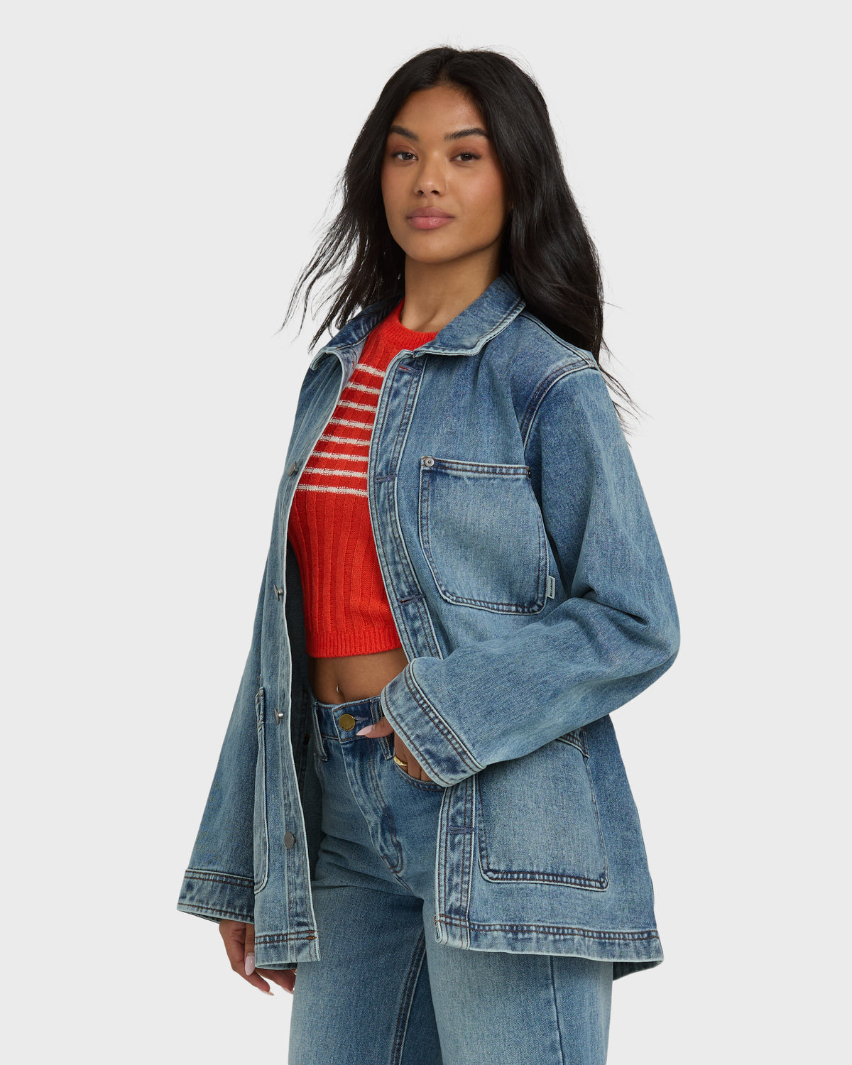 Medium-wash denim chore coat with collar, button front, and patch pockets worn over red striped shirt and matching jeans.