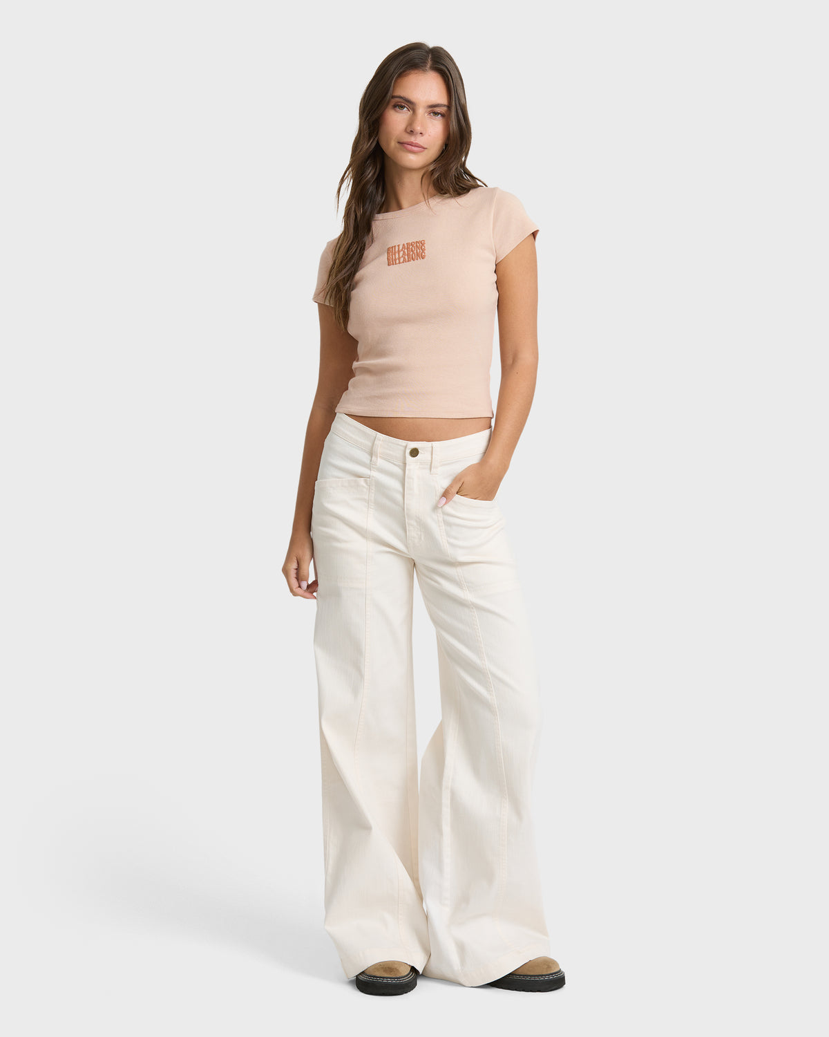 Oatmeal short sleeve t-shirt with small orange text graphic on chest, shown on model with white jeans.