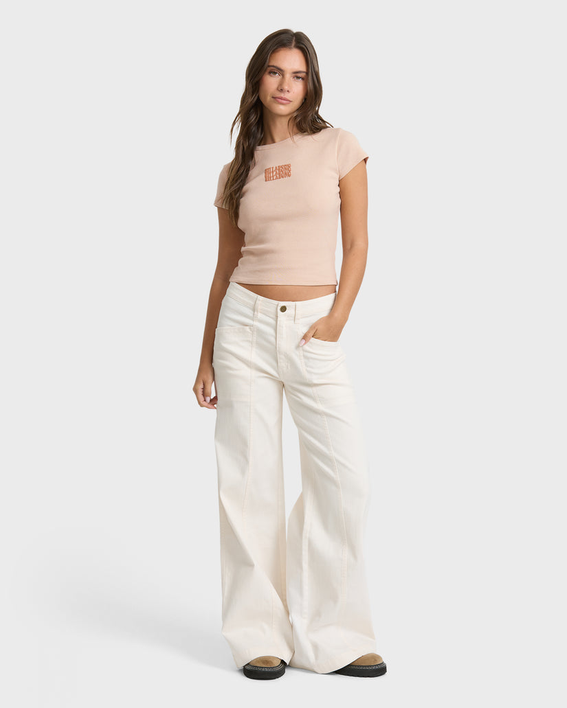 Oatmeal short sleeve t-shirt with small orange text graphic on chest, shown on model with white jeans.