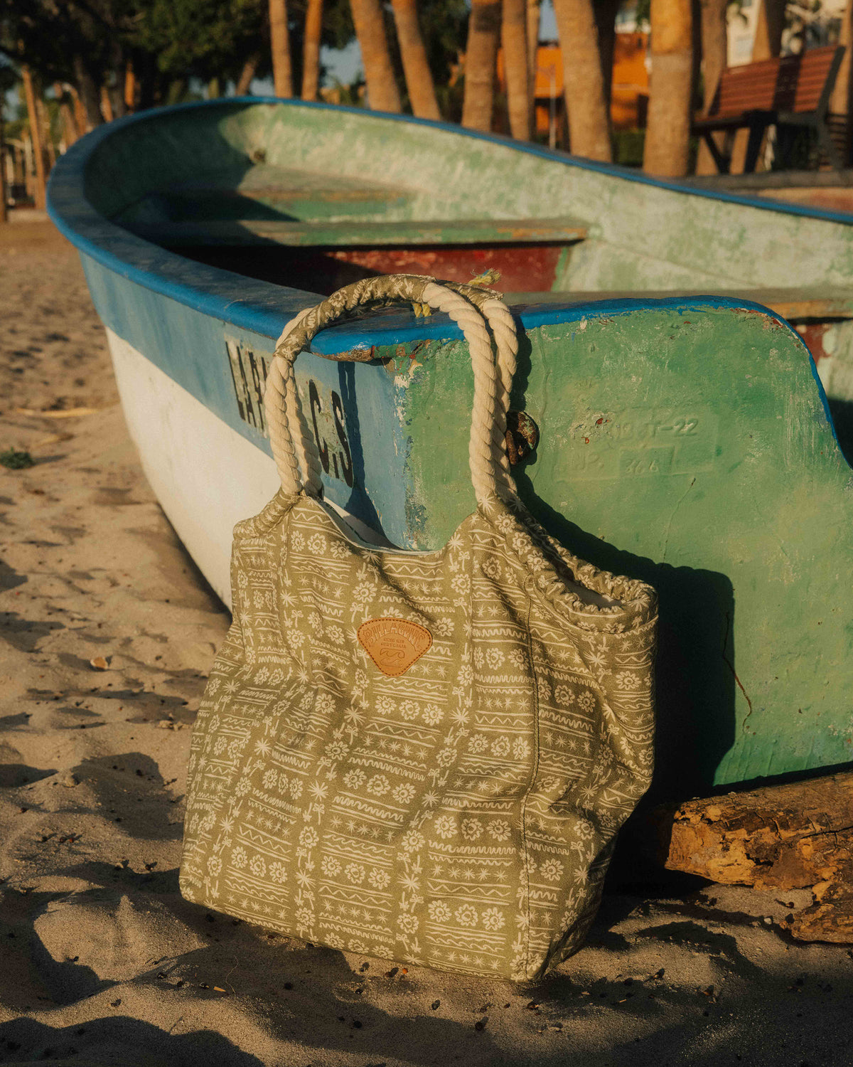 Green tote bag with rope handles and geometric pattern, displayed on beach sand next to boat.