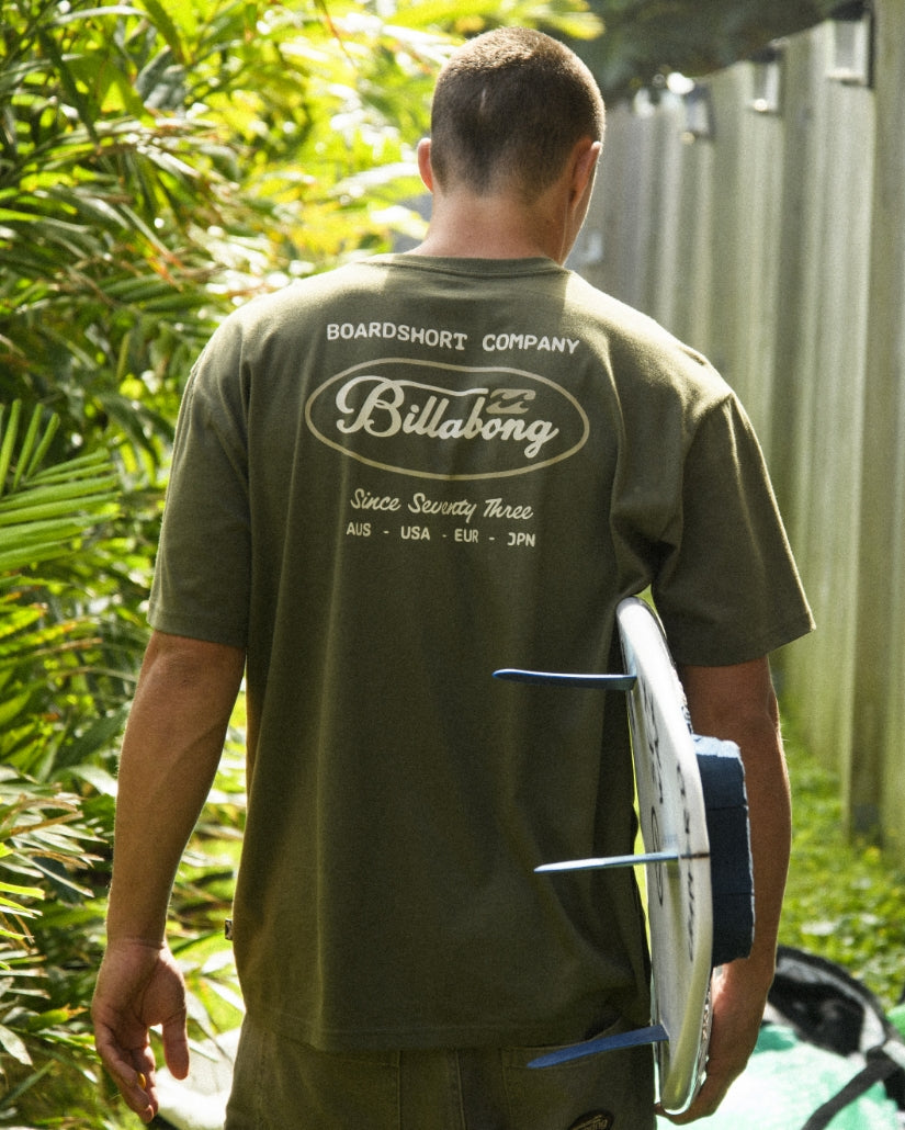 Olive short-sleeve tee with "Boardshort Company" and "Billabong" text printed on back, worn by man holding surfboard.