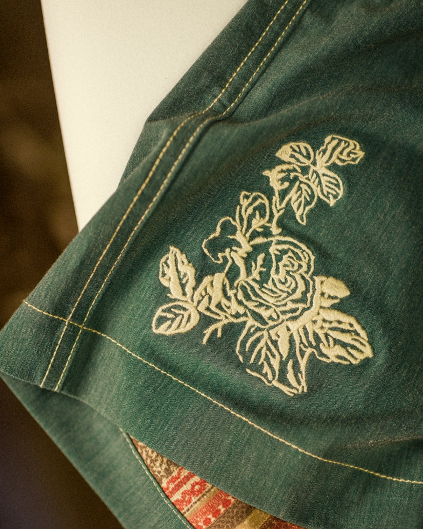 Teal boardshorts with cream-colored floral embroidery on front and yellow topstitching detail.