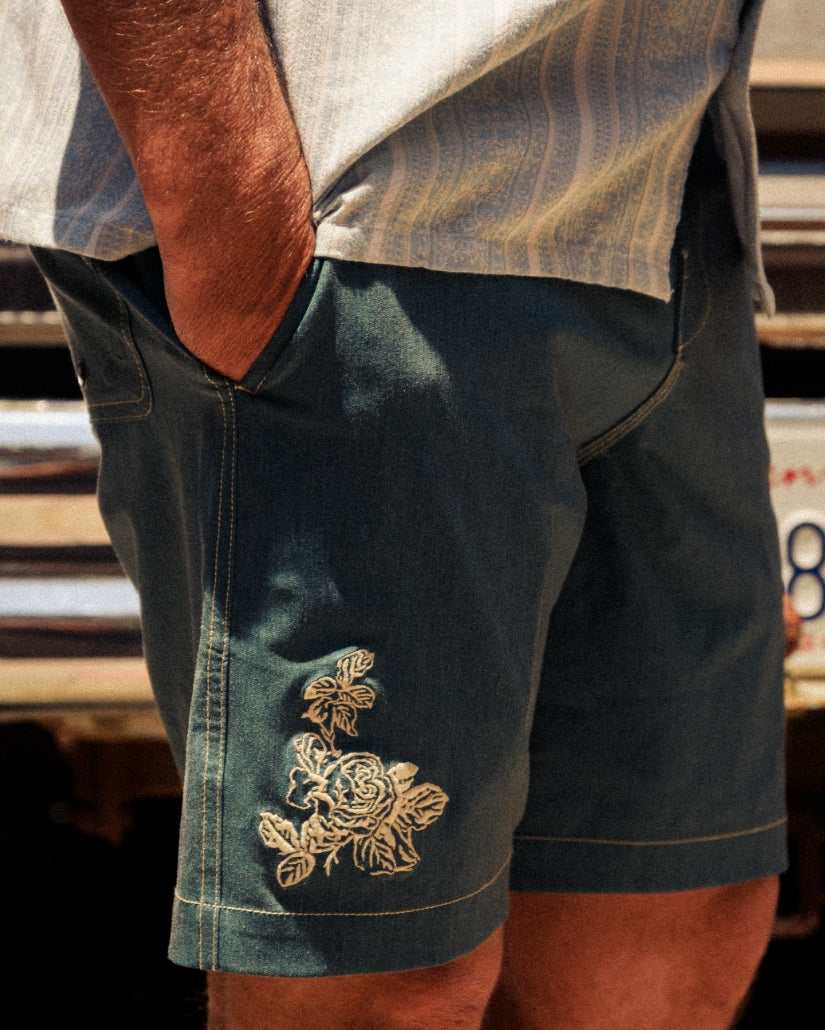 Teal boardshorts with gold floral embroidery on the leg, worn with a light-colored shirt.