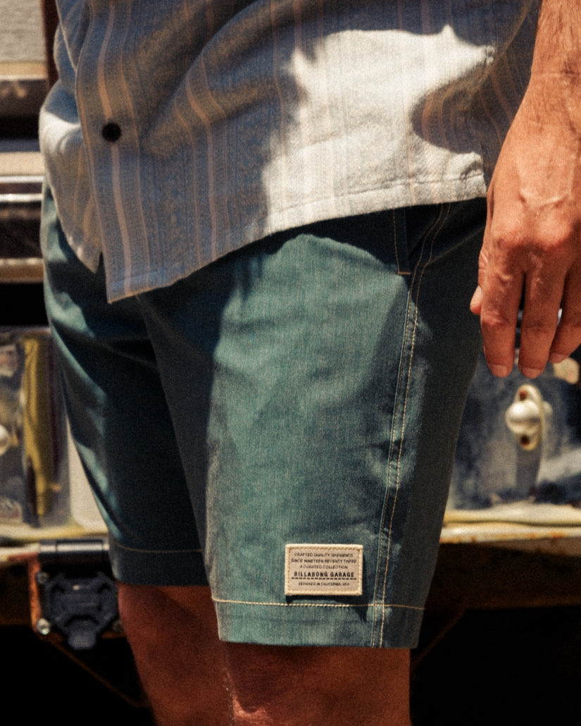 Teal boardshorts with black side panels and white waistband, worn with striped shirt.
