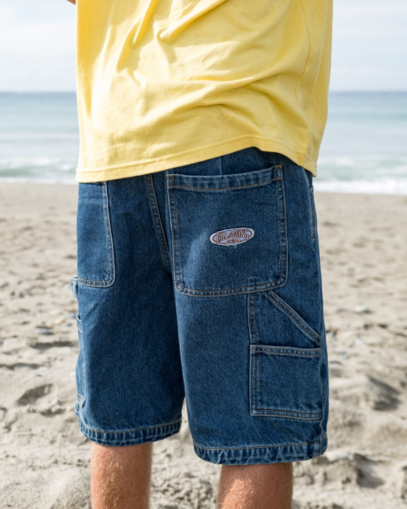 Dark blue denim cargo shorts with multiple pockets and Billabong logo patch, worn on beach.