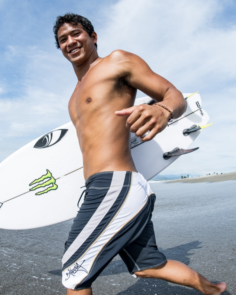 Man wearing dark navy and white striped boardshorts with yellow accent stripe, holding a surfboard at the beach.