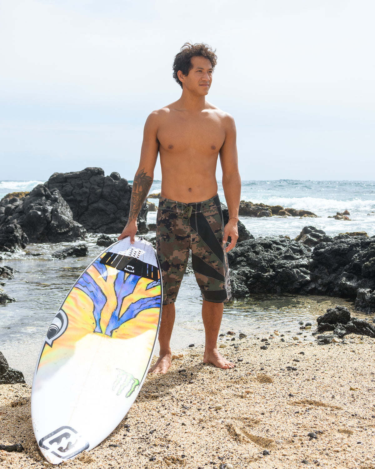 Man wearing dark olive camouflage board shorts holds a yellow and blue surfboard on a beach.