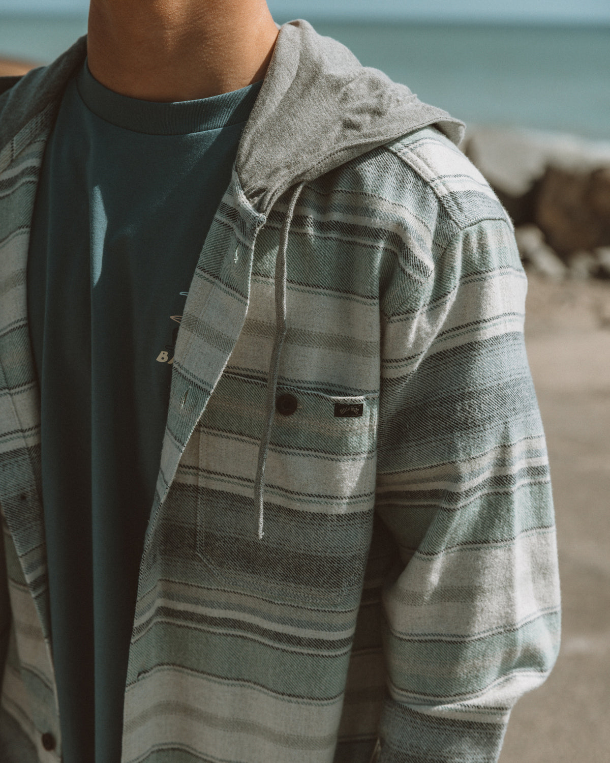 Oatmeal flannel shirt with green and cream horizontal stripes, hood, and drawstring worn over dark t-shirt.