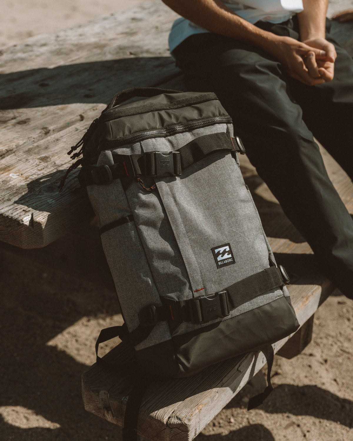 Heather grey backpack with dark straps and zippered compartments, sitting on wooden surface next to person.