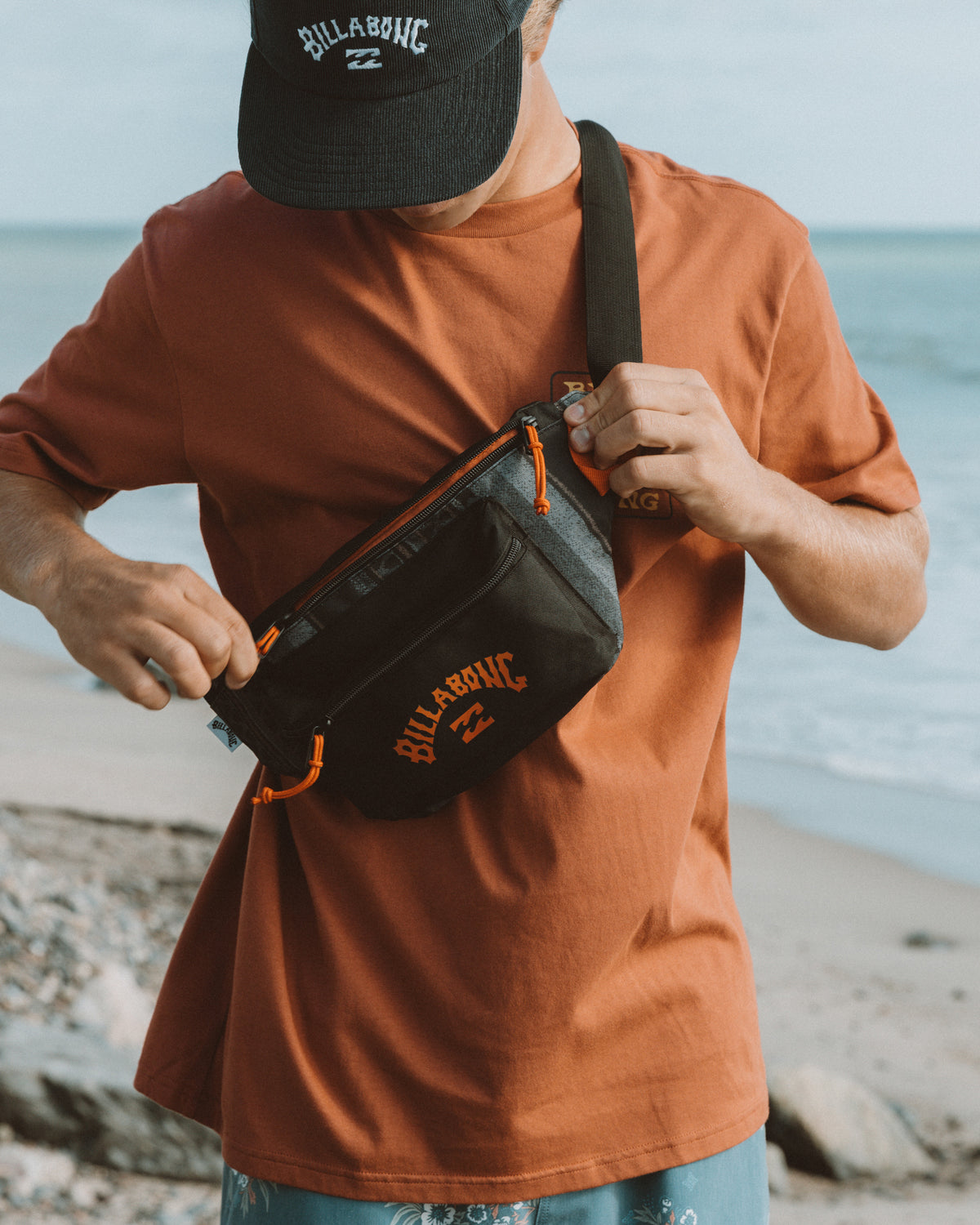 Black and grey backpack with orange accents and "Billabong" branding, worn as crossbody bag at the beach.