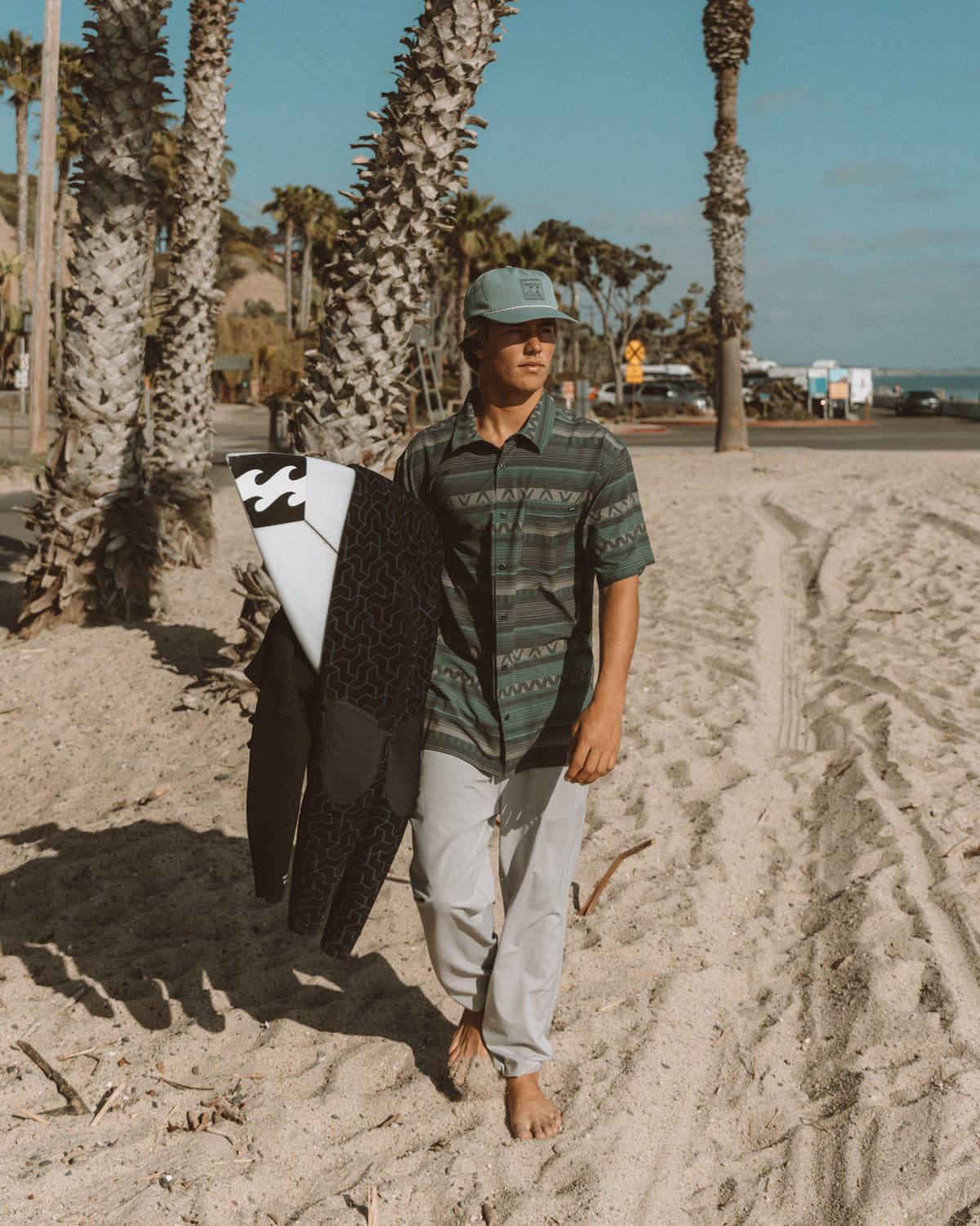 Man wearing gray Surftrek Transit Joggers with striped shirt, holding surfboard on beach with palm trees.
