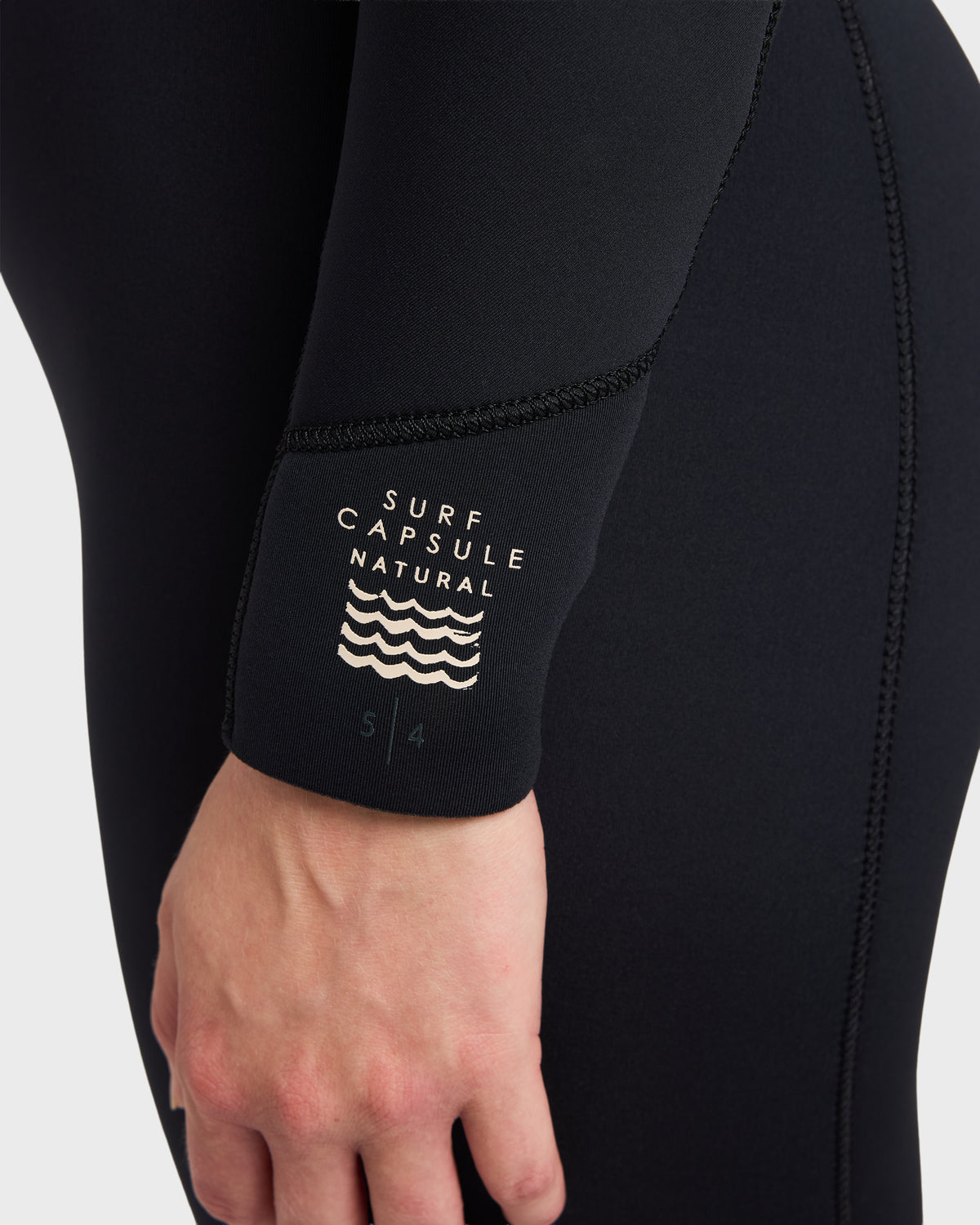 Black full wetsuit with chest zip, taped seams, and "Surf Capsule Natural" label on wrist.