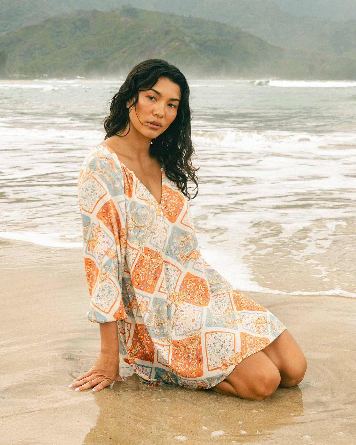 Woman wearing white mini dress with orange and blue patterned design, kneeling on beach with ocean waves in background.