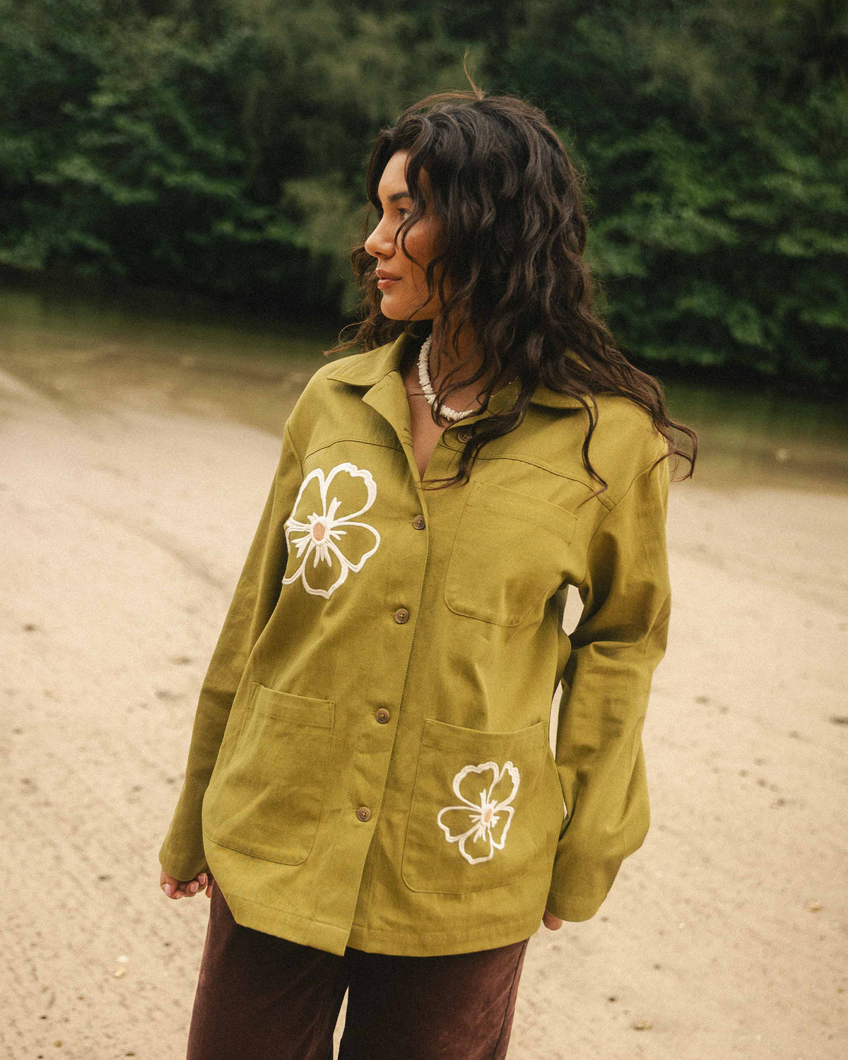 Moss-colored shacket with white floral embroidery on chest and pocket, button front, worn over brown pants.