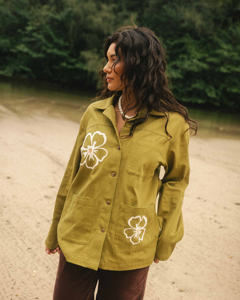 Moss-colored shacket with white floral embroidery on chest and pocket, button front, worn over brown pants.