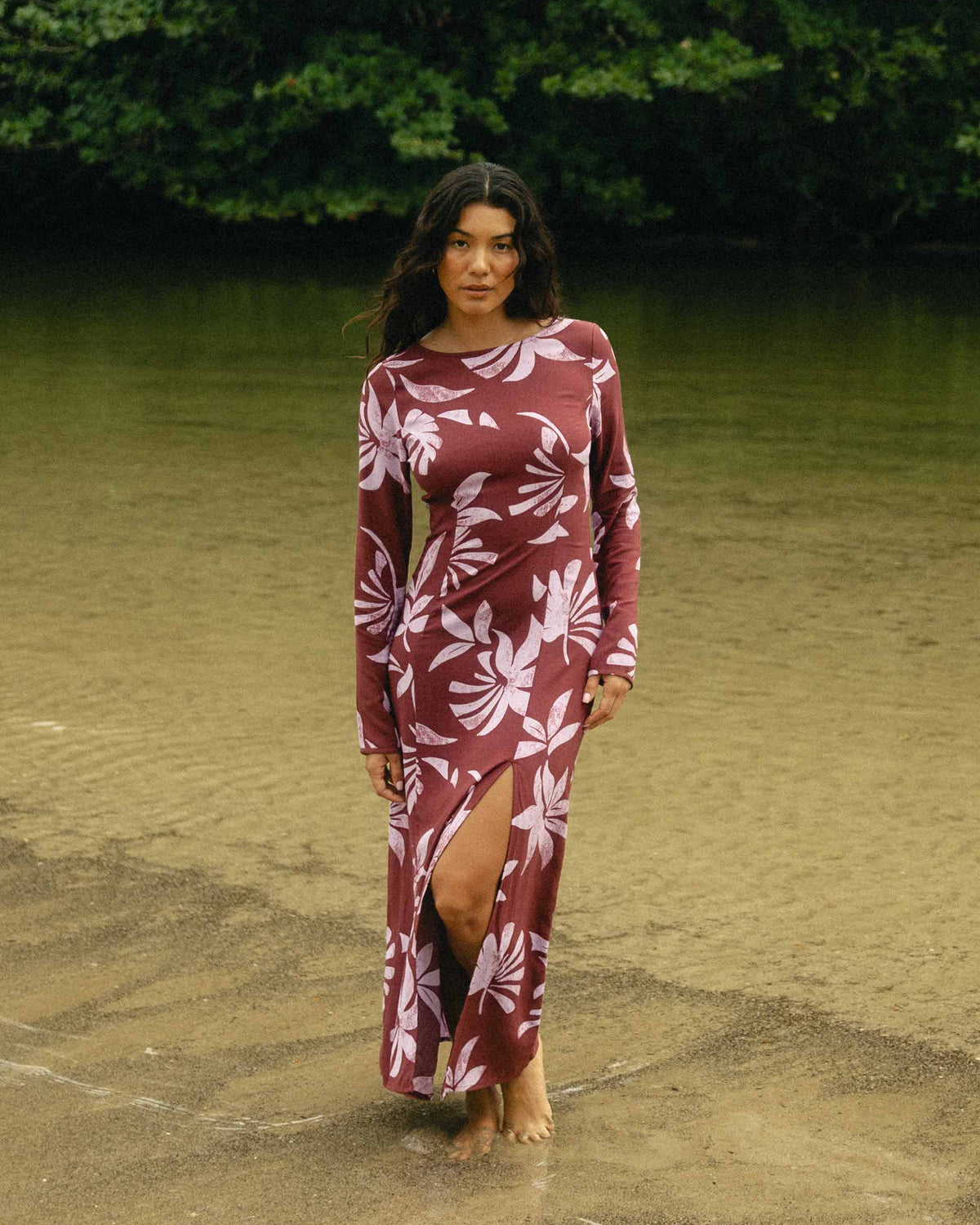Burgundy maxi dress with white floral print, long sleeves, and side slit, worn by woman standing barefoot outdoors.