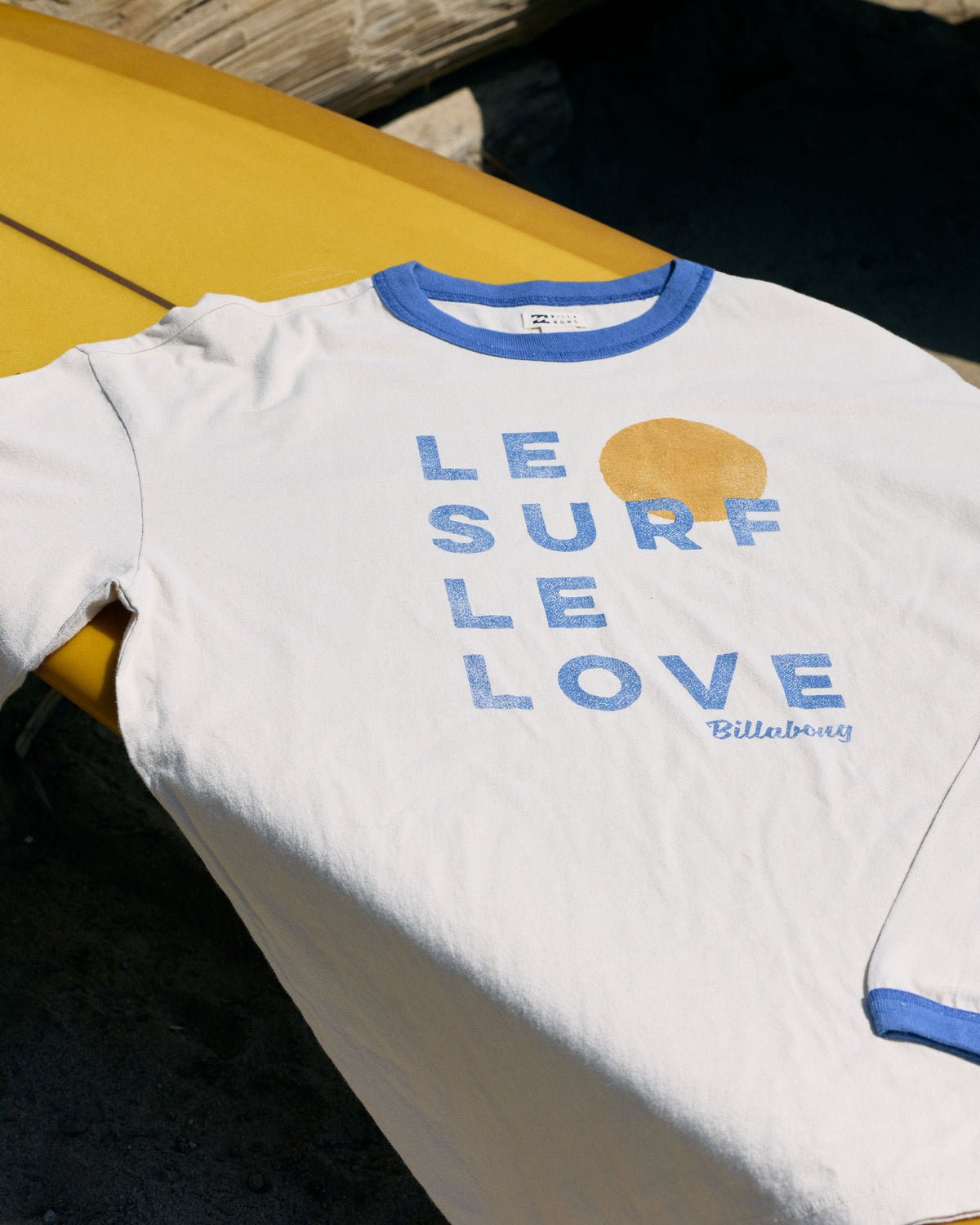 White long sleeve ringer t-shirt with blue trim and "Le Surf Le Love" text with gold sun graphic.