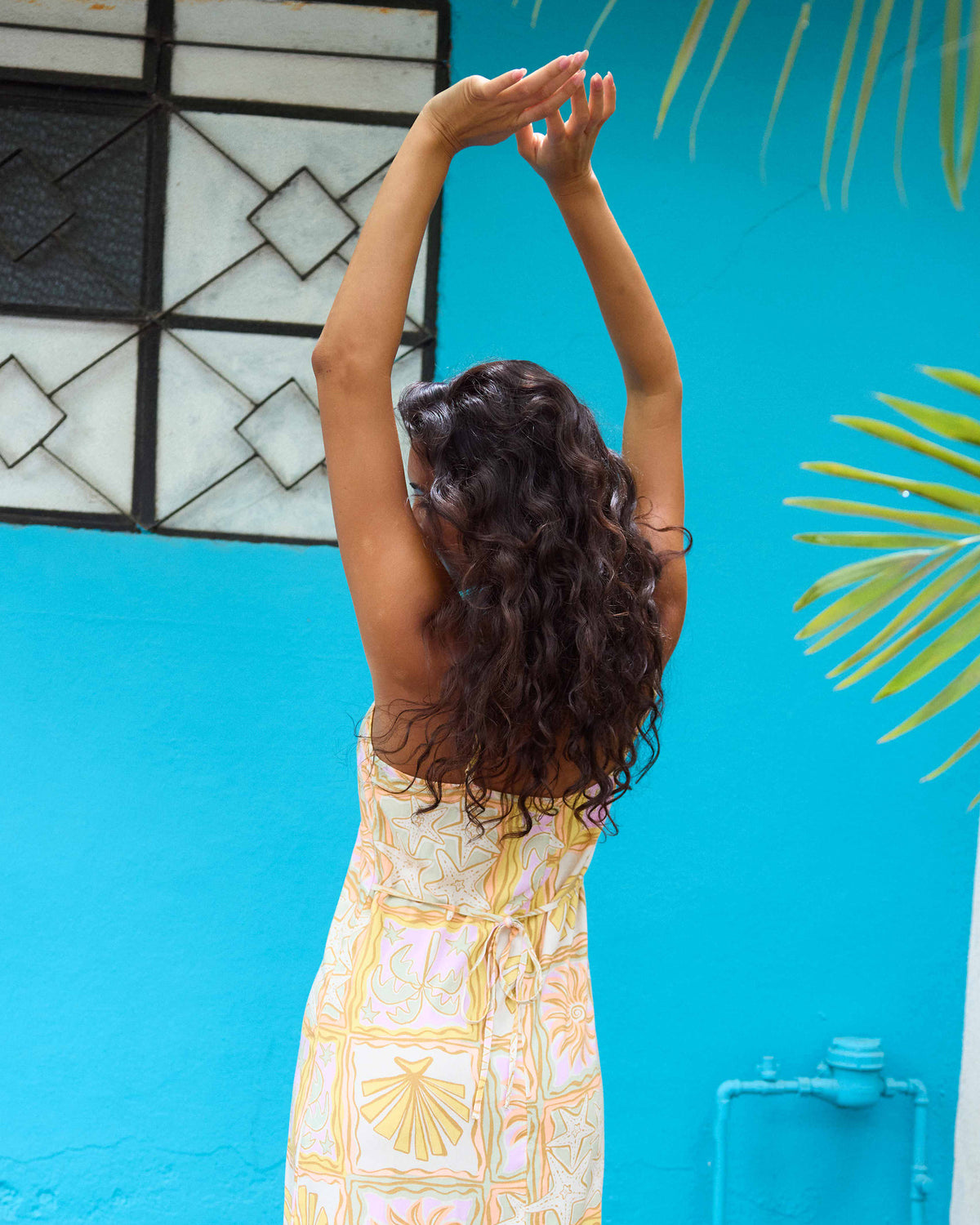Woman wearing lilac midi dress with yellow and white geometric print, photographed from behind against blue wall.