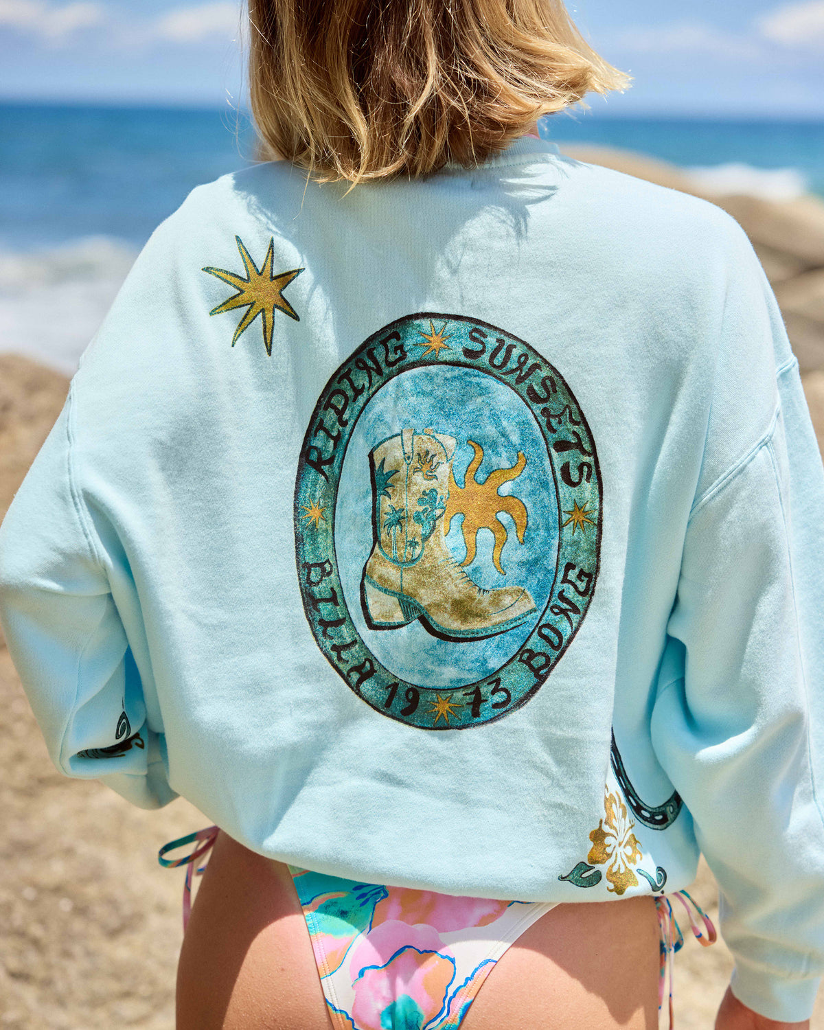 Clearwater crewneck sweatshirt with sun and surfboard graphic on back, worn at beach.