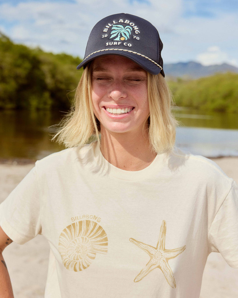 White short sleeve t-shirt with gold shell and starfish graphics, styled with a navy baseball cap.