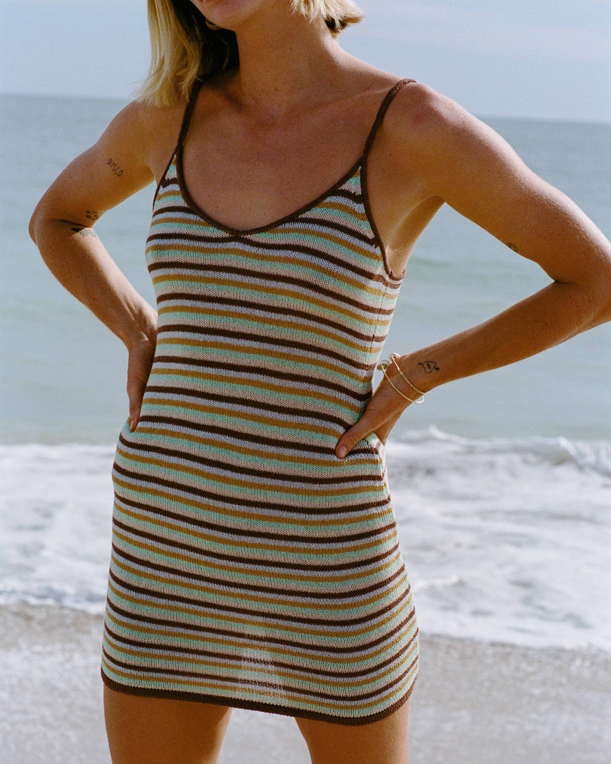 Woman wearing brown, tan, and green striped mini dress with thin straps at the beach.