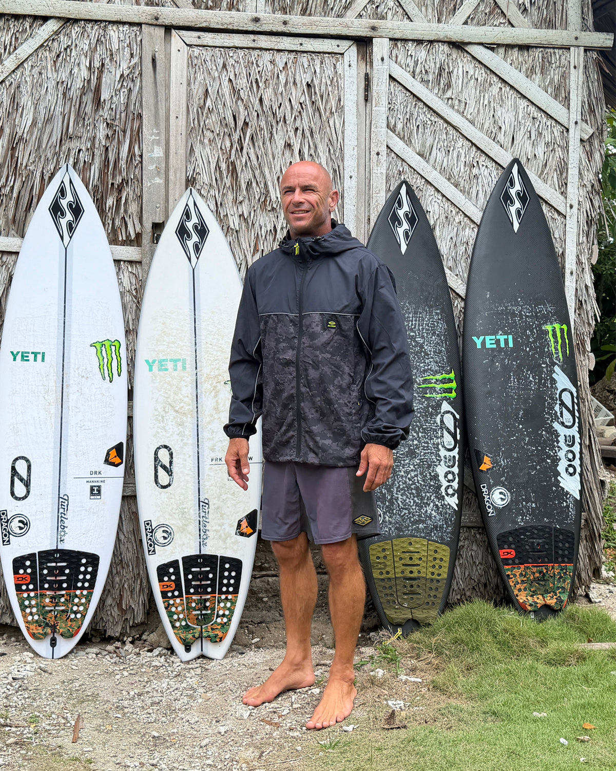 
A barefoot man in a black hooded windbreaker and gray shorts stands before four surfboards leaning against a thatched wall.