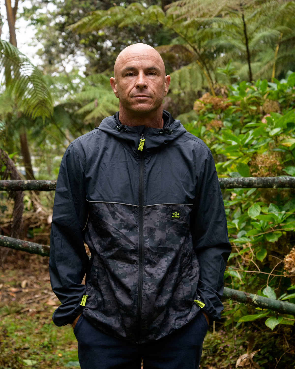 
A bald man stands outdoors among ferns wearing a black hooded zip jacket with a gray digital camouflage lower half and neon yellow zipper pulls.