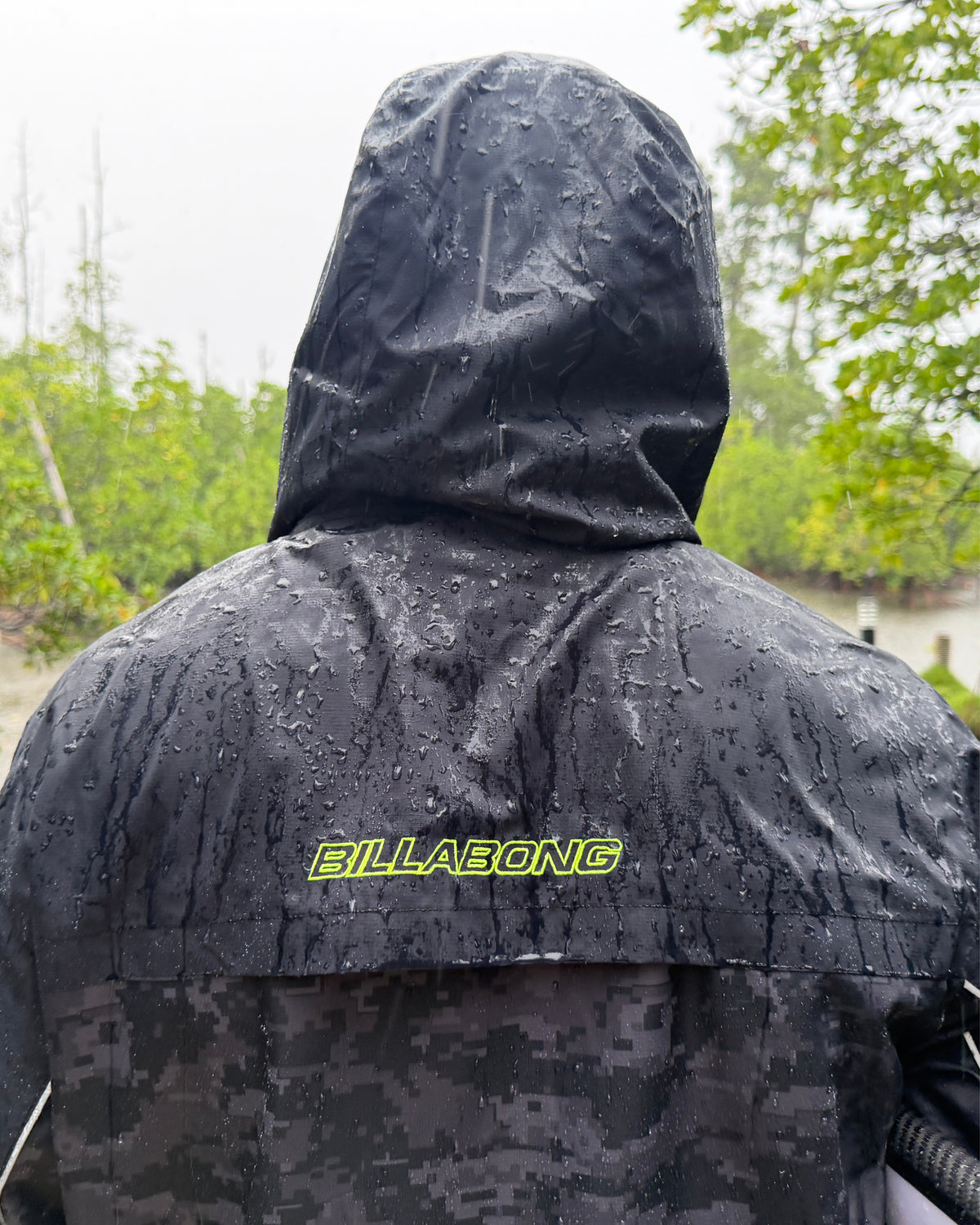 
Rear view of a person wearing a black hooded jacket covered in raindrops, with a neon yellow Billabong logo and digital camouflage panel.