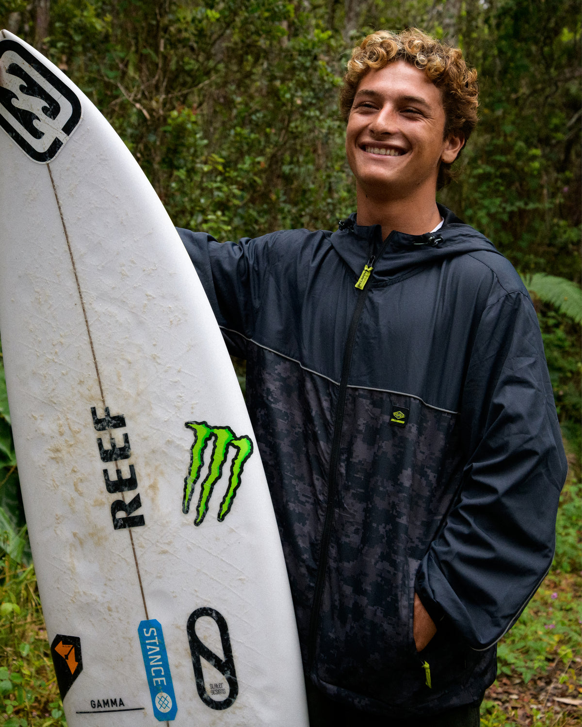 
A smiling young man with curly hair wears a dark hooded windbreaker with a camouflage lower panel while holding a white surfboard outdoors.