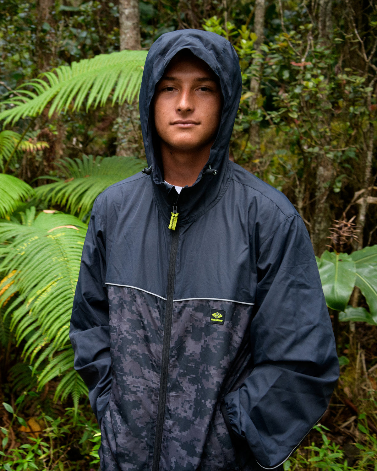 
A young man wears a dark hooded windbreaker with a digital camouflage lower panel and a lime zipper pull, standing among ferns.