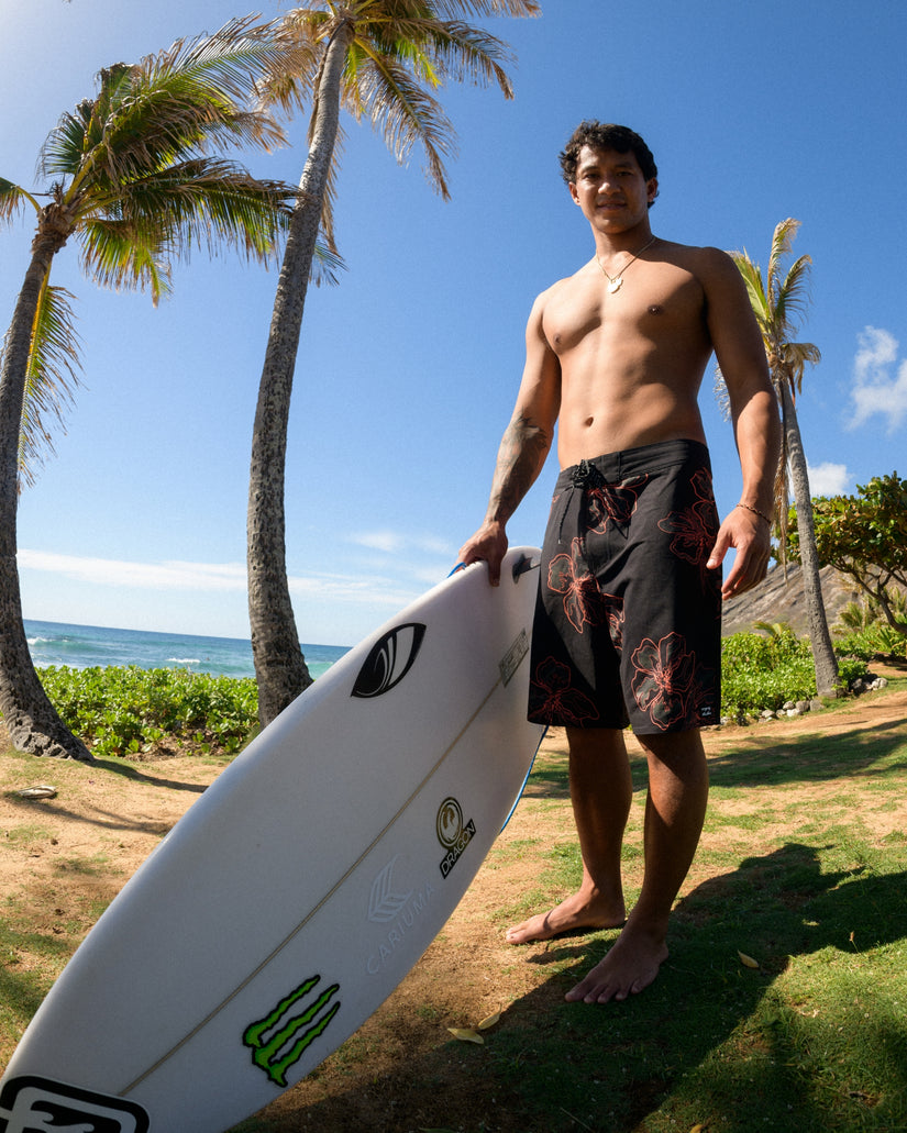 Black boardshorts with red and orange floral print, 19" inseam, worn by man holding surfboard on beach.