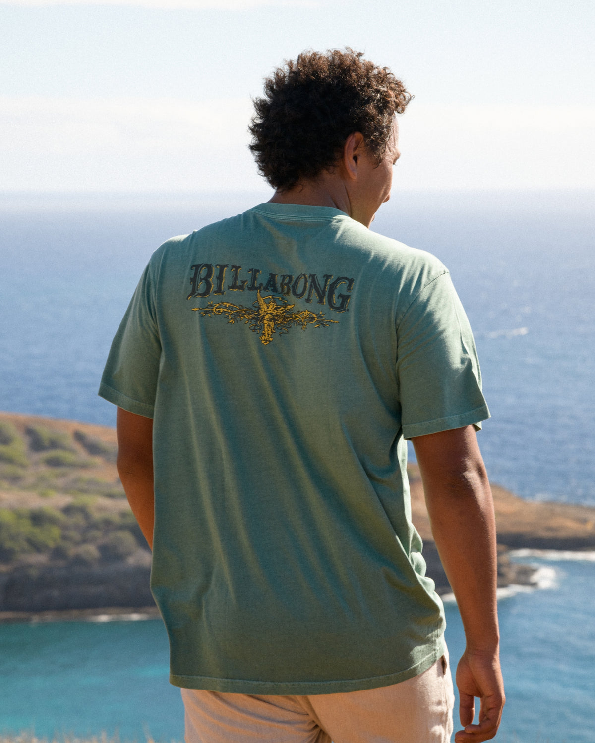 Sea green short sleeve t-shirt with "Billabong" text and graphic design on back, worn by person facing ocean.