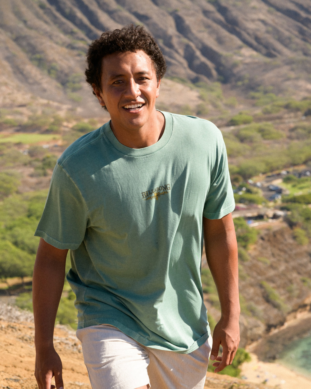 Sea green short-sleeve t-shirt with small chest logo, worn by man with white shorts against mountain landscape.
