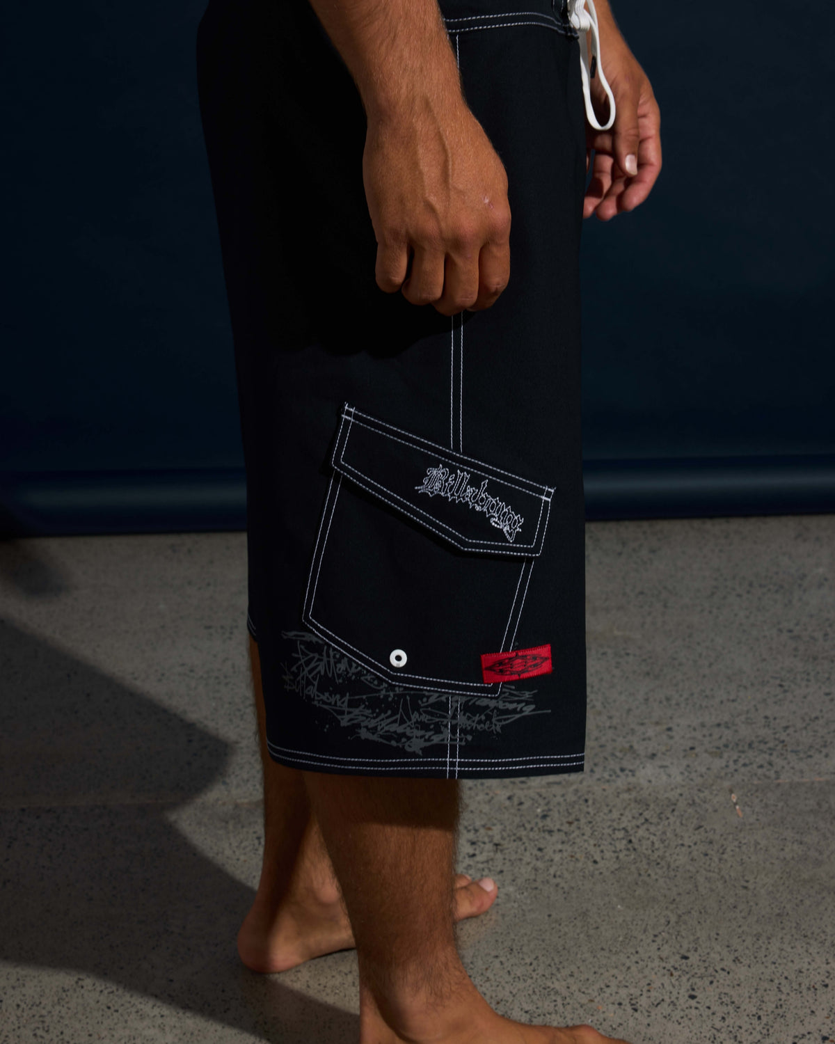 Black boardshorts with white stitching, chest pocket, and red label detail worn by a man.