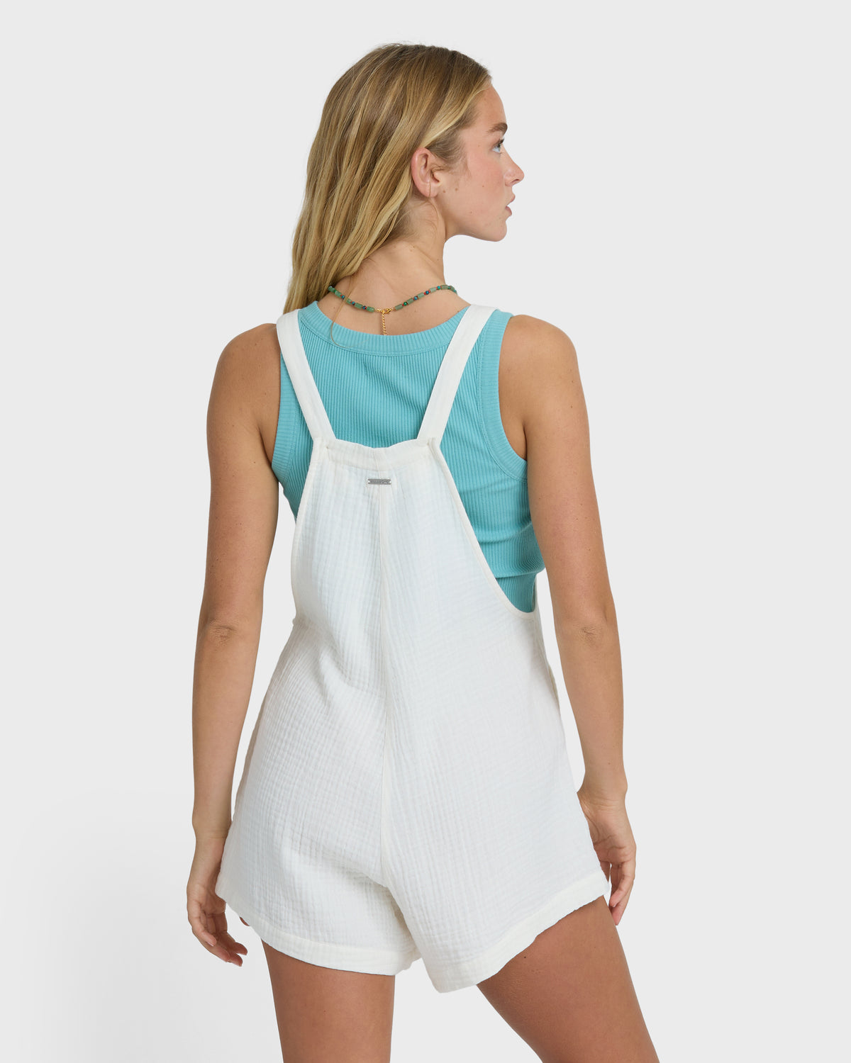 Woman wearing white textured romper with turquoise ribbed tank top underneath, shown from back view.