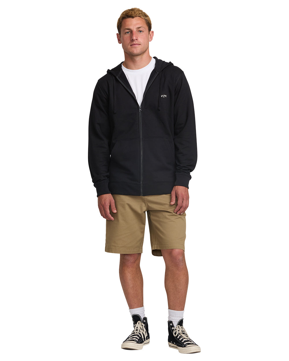 Man wearing black zip-front hoodie with hood, white undershirt, tan shorts, and black canvas sneakers.