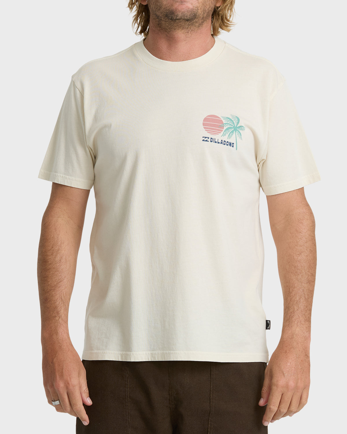 
A man wears an oatmeal short sleeve t-shirt with a small pink sun, teal palm tree, and Billabong logo on the chest.